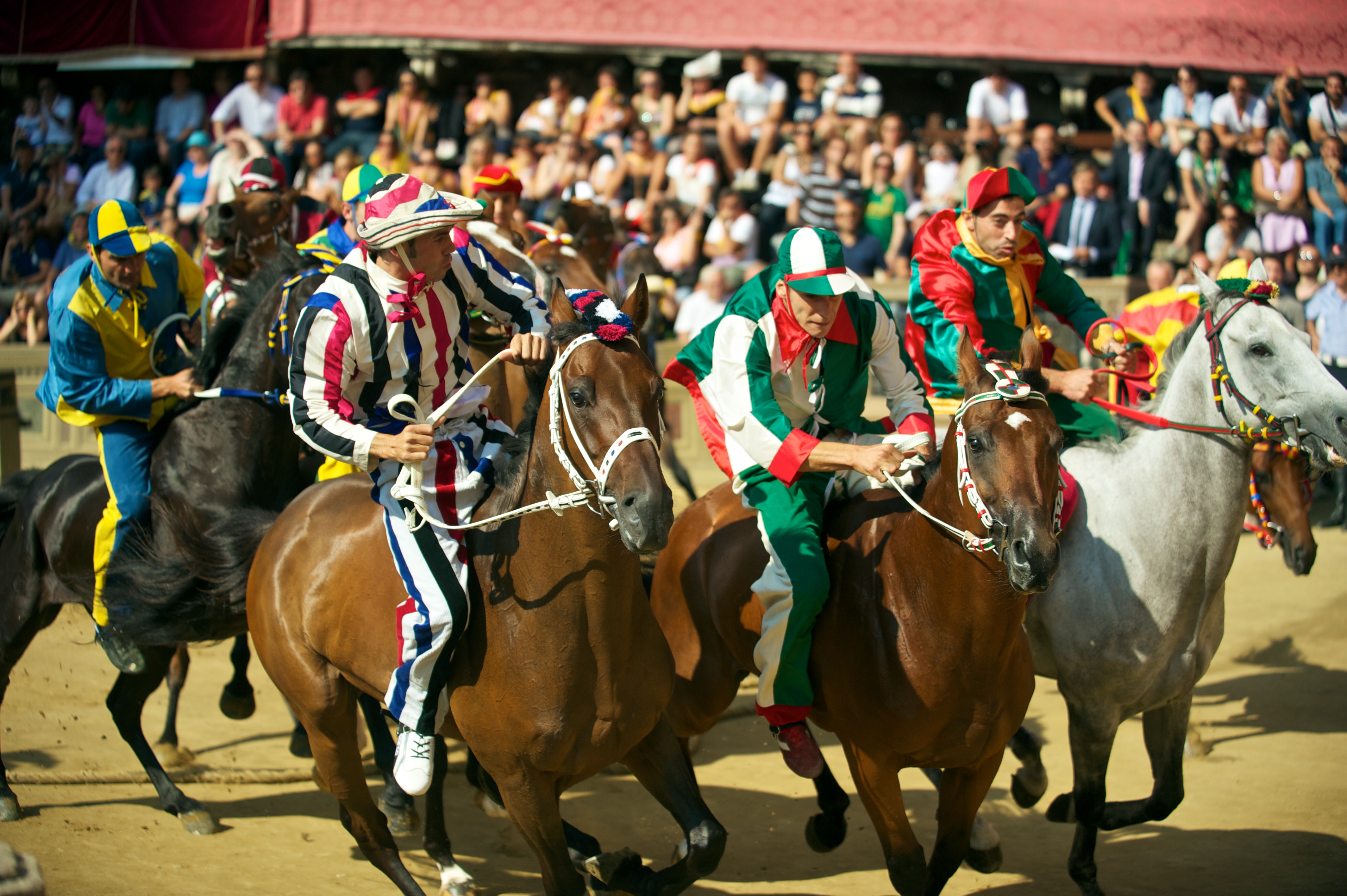 Colorfully costumed jockeys race bareback on horseback during the Palio di Siena, the historic medieval horse race held in Siena's Piazza del Campo, Italy, with a packed crowd of spectators in the background.